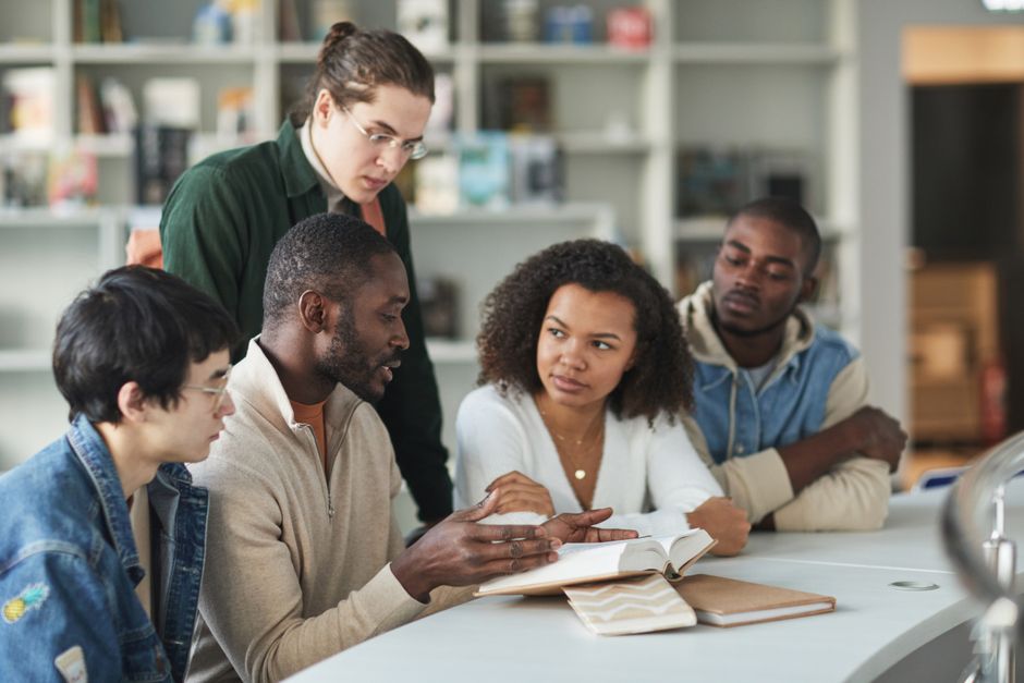 A diverse group of college students discussing a book in a library setting, showcasing teamwork and education.